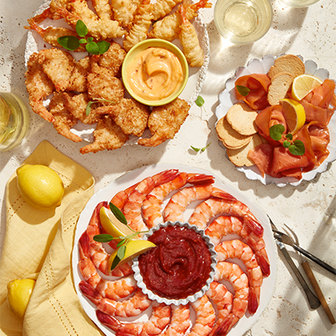 Seafood appetizer spread with a platter of shrimp and cocktail sauce, a plate of coconut shrimp with dipping sauce, and smoked salmon with crackers and lemon slices, served with glasses of white wine and fresh lemons.
